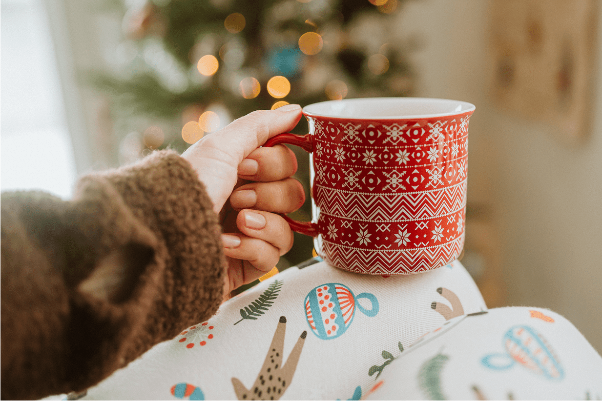 Hand holding festive mug near Christmas tree.